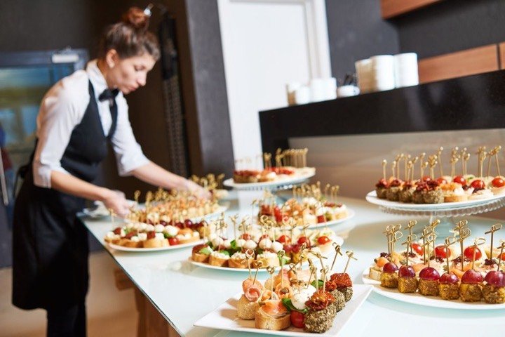 A catering server in an apron arranging platters of hors d'oeuvres on a buffet table